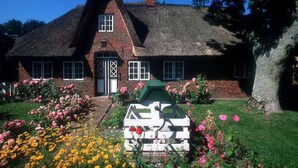 Ein traditionelles Haus mit Strohdach und Backsteinfassade, einer Zwerchhaus-Tür mit Diamantmusterfenster und einem kleinen Garten mit Blumen und einer weißen Bank.