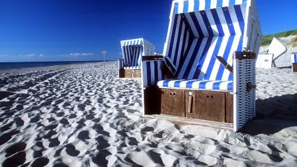 Zwei gestreifte Liegestühle an einem sandigen Strand unter blauem Himmel.