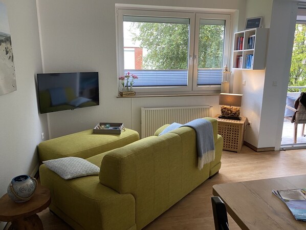 A living room with a sofa, a TV, a radiator, a window with blinds, a lamp, and a bookshelf. - Apartment 'Felix, 35qm' with Balcony (Horumersiel-Schillig)