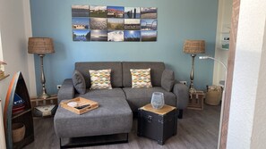 Un salon avec un canapé gris, deux coussins à motifs, une table d’appoint en bois, une lampe et un collage mural.