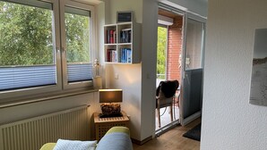 Een kamer met een bank, een klein tafeltje met een lamp, een boekenkast en uitzicht op het omliggende landschap door een schuifdeur.