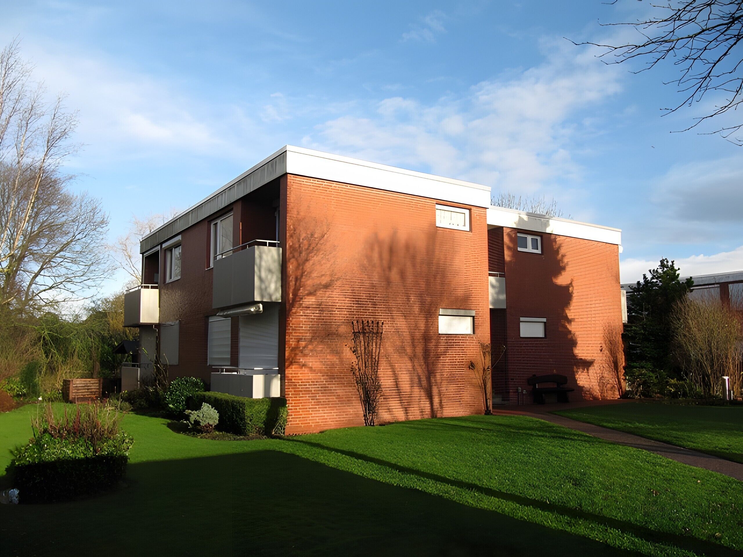 Ein zweigeschossiges Backsteingebäude mit flachem Dach, einem kleinen Balkon im Erdgeschoss und einem gepflegten Rasen.