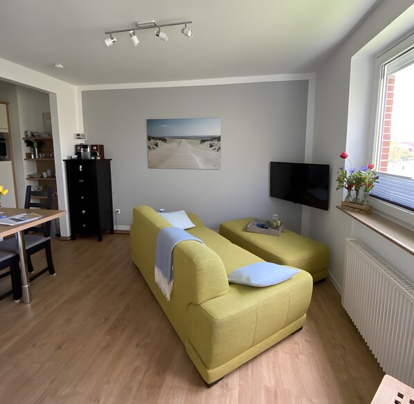 Una sala moderna con sofá, televisor, comedor con mesa y sillas, y área de cocina con armarios y refrigerador.