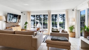 A modern living room with a sofa, armchair, and ottoman, a flat-screen TV mounted on the wall, and large windows with curtains.