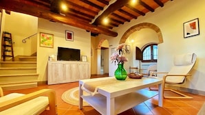 A living room with a white sofa, a wooden bench, a dining table with a vase, and a TV on a cabinet. - Montagnana Country House - Relax and stunning view (San Gimignano)