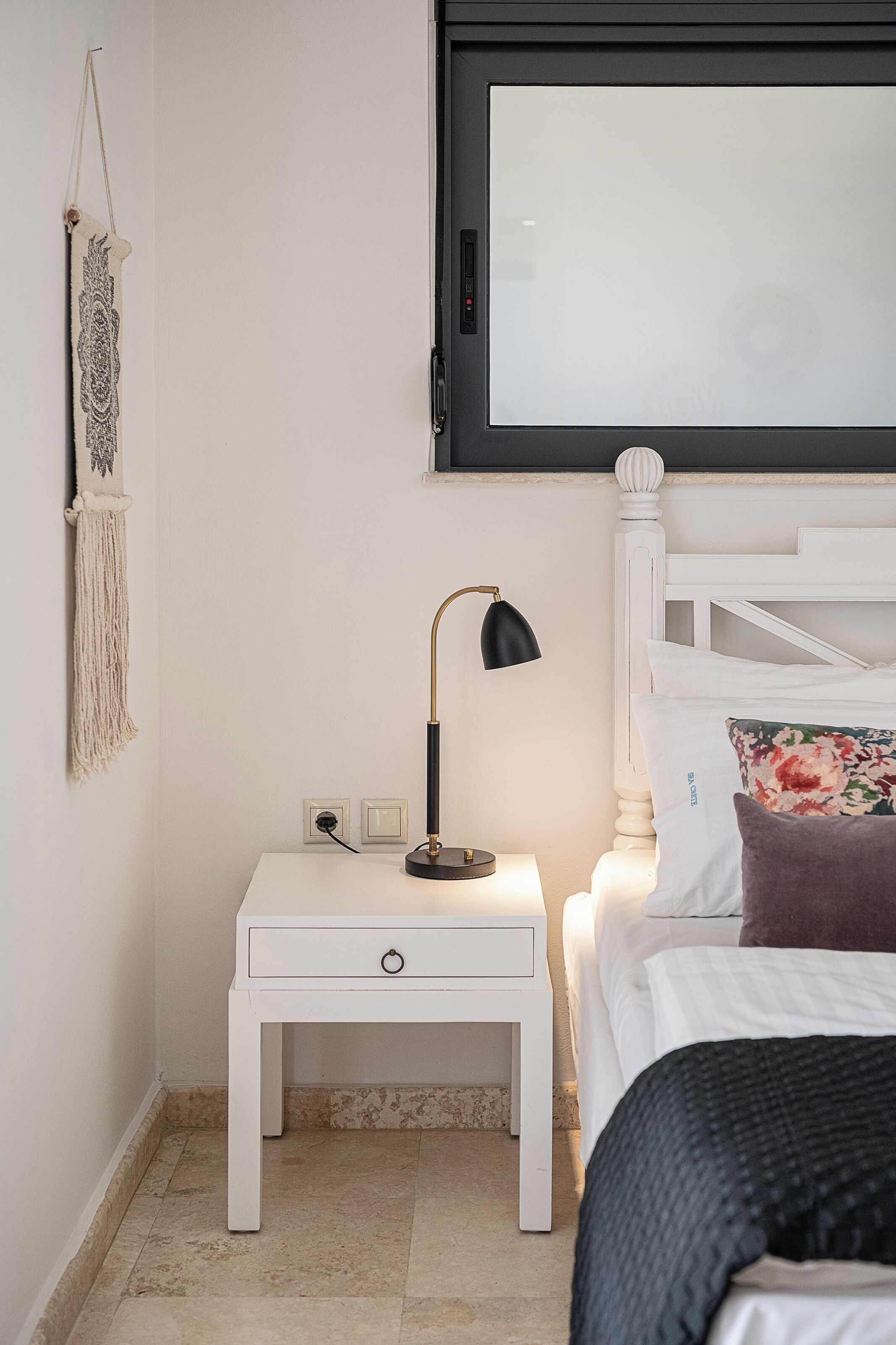Une table de chevet blanche avec une lampe, un lit avec des oreillers blancs à motifs floraux et une décoration murale en macramé.