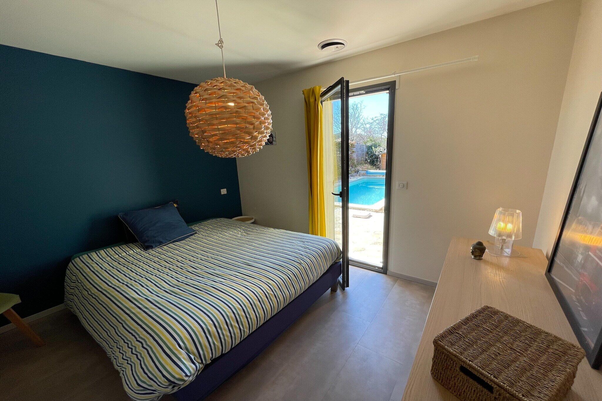 Une chambre avec un lit, un luminaire suspendu, une petite table avec une lampe et une vue sur une piscine, visible à travers une porte vitrée.