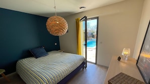 Ein Schlafzimmer mit einem Bett, einer Hängeleuchte, einem kleinen Tisch mit Lampe und Blick auf einen Swimmingpool durch eine Glasdoor.