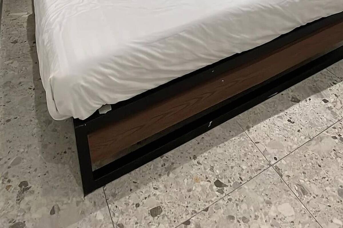 Een slaapkamer met een bed, houten hoofdbord, nachtkastje, raam met jaloezieën en een wandlamp.