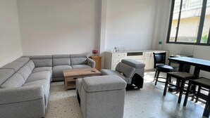 Una sala moderna con un sofá seccional gris, una mesa de centro de madera y un pequeño otoman. Se observa un escritorio con una silla y un ventanal que proporciona luz natural.