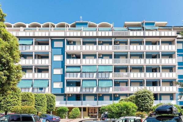 A multi-story hotel with balconies, air conditioning units, and a parking area in front.