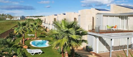 A modern hotel with a swimming pool, palm trees, and a covered outdoor area with lounge chairs.
