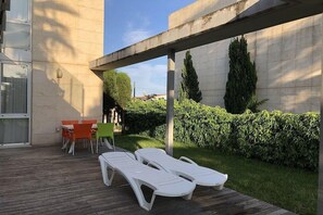 Um pátio com cadeiras de descanso e uma mesa, cercado por um arbusto e um edifício.