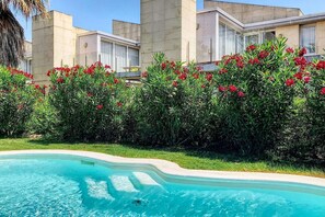 A swimming pool surrounded by lush greenery and red flowers, with modern buildings in the background.