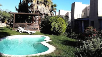 Una piscina con muelle de madera y sillas de descanso, rodeada de follaje y una palmera.