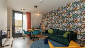 Una sala moderna con un sofá verde, un sillón amarillo y un tapete azul. Hay una zona de comedor con una mesa y sillas, y un ventanal con cortinas.