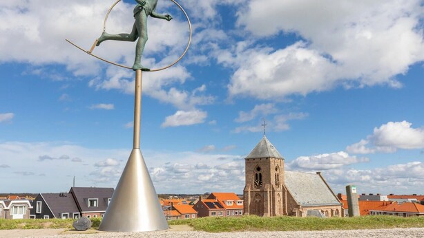 Una escultura de una figura equilibrándose sobre un palo con un aro, frente a un fondo de casas y una iglesia.