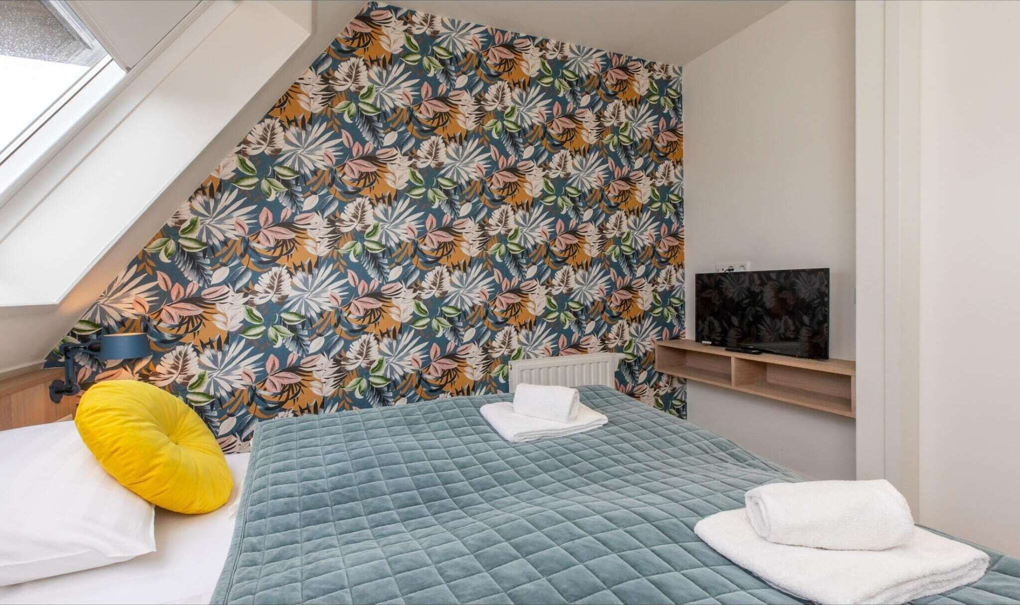 A bedroom with a sloped ceiling, a bed with a quilted blue cover, a yellow pillow, a white pillow, a folded white towel, and a TV on a wooden shelf.