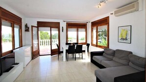 Ein modernes Wohnzimmer mit einem grauen Sofa, einem Esstisch mit Stühlen und einem Fernseher auf einem Schrank. Große Fenster mit Holzrahmen, und durch die Glastüren ist ein Balkon zu sehen.