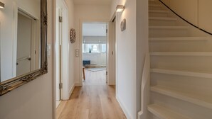 A hallway with a wooden floor, a white staircase, and a door leading to a room with a window and a plant.