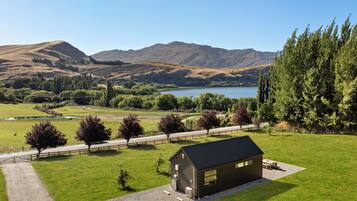 Una cabaña rústica con camino de grava, rodeada de abundante vegetación y un lago, con montañas al fondo.