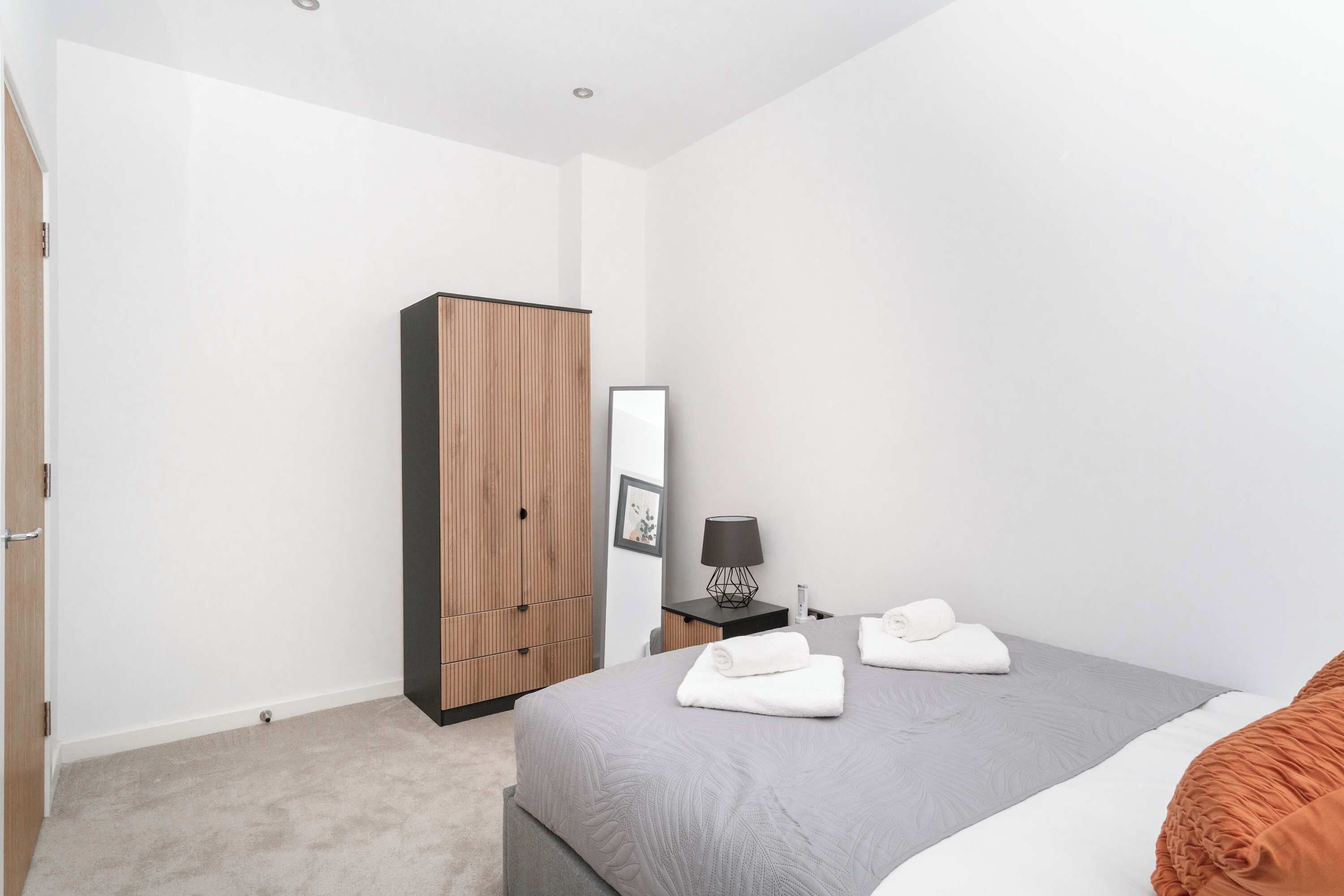 A modern bedroom with a grey bed, a wooden wardrobe, a bedside table with a lamp, and a framed picture on the wall.
