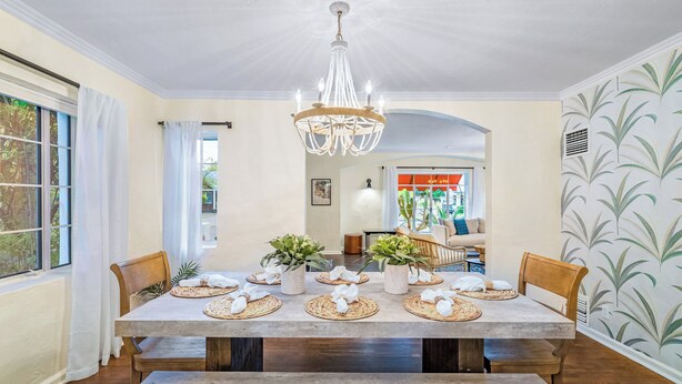 A dining room with a large table set for six, surrounded by chairs, a chandelier, and potted plants.