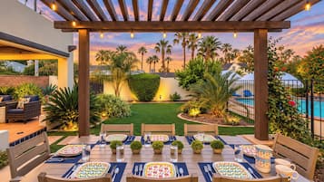 Un patio exterior con una pérgola de madera, mesas de comedor preparadas para una comida, y una piscina al fondo.
