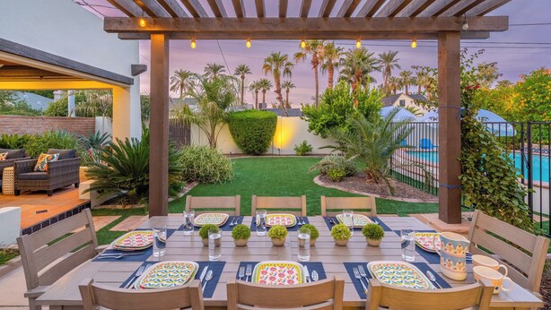 An outdoor dining area with a pergola, tables set for four, and a view of a pool and palm trees. - Timeless Elegance: Historic Estate w/ Pool, Hot Tub + Putting Green (Phoenix)