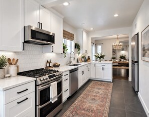 A modern kitchen with white cabinetry, a gas stove, a built-in oven, a microwave, and a dishwasher. There is a large window with a view of the outdoors, and a patterned rug on the floor. - Timeless Elegance: Historic Estate w/ Pool, Hot Tub + Putting Green (Phoenix)