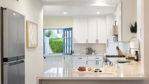 A modern kitchen with white cabinets, a stainless steel refrigerator, a large window with a view of greenery, and a countertop with fruits and a coffee maker. - Timeless Elegance: Historic Estate w/ Pool, Hot Tub + Putting Green (Phoenix)