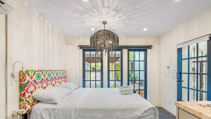 A bedroom with a bed, a chandelier, a colorful headboard, and a view through blue doors.