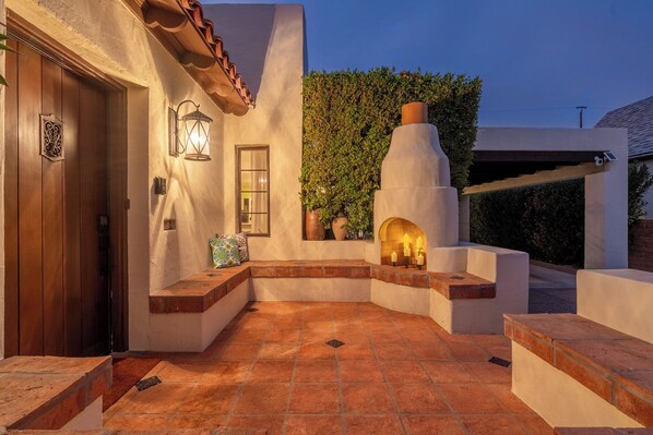 A courtyard with a stone floor, a wooden door, a wall-mounted lantern, and a brick oven.