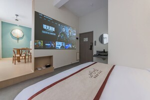 Habitación de hotel con cama, televisor mostrando una película, una silla de madera, una mesita y un estante con pantuflas.