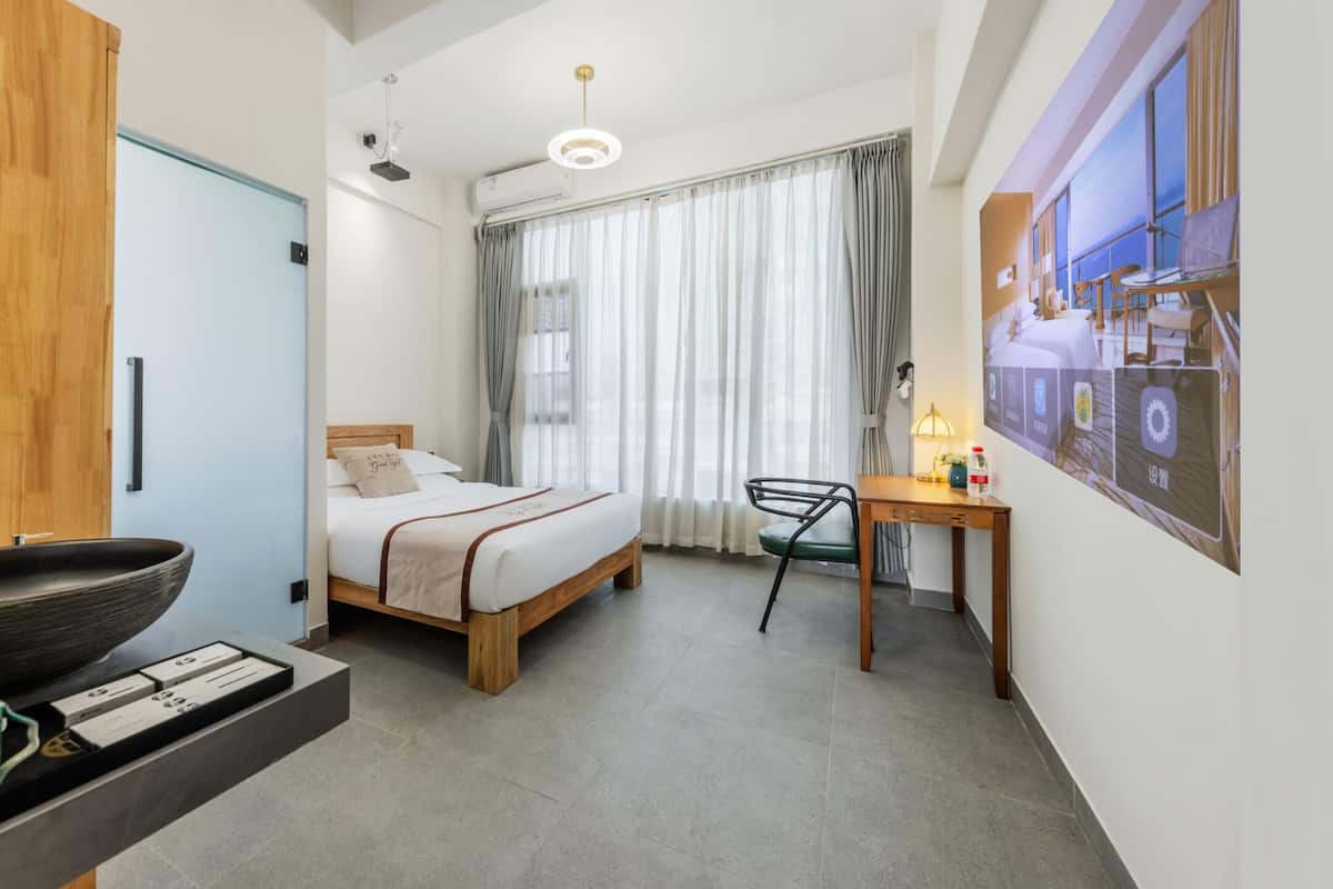 A hotel room with a bed, a desk, a chair, a window with curtains, and a wall-mounted air conditioner.