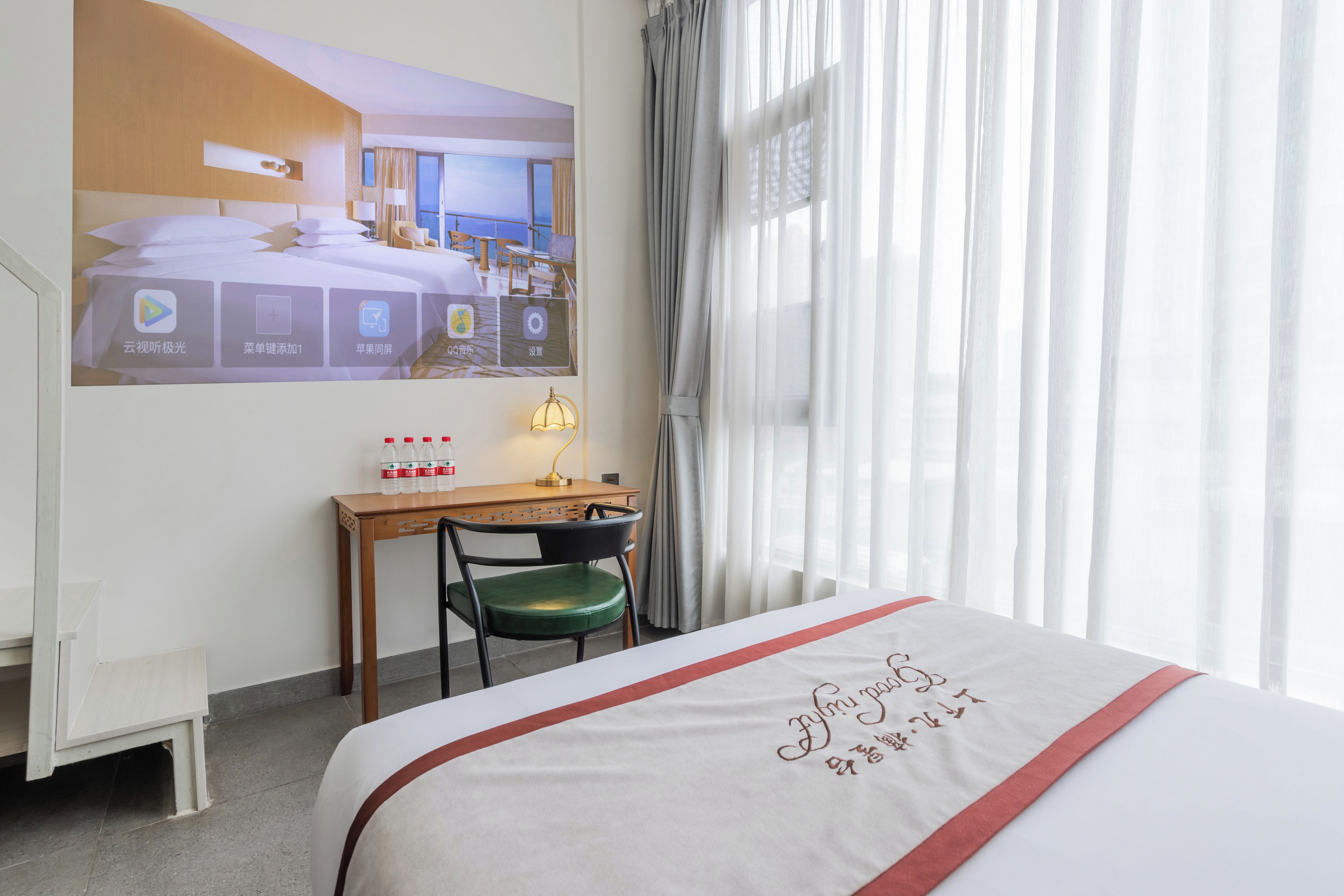 A hotel room with a bed, a desk with a chair, a small table, and a window with curtains.