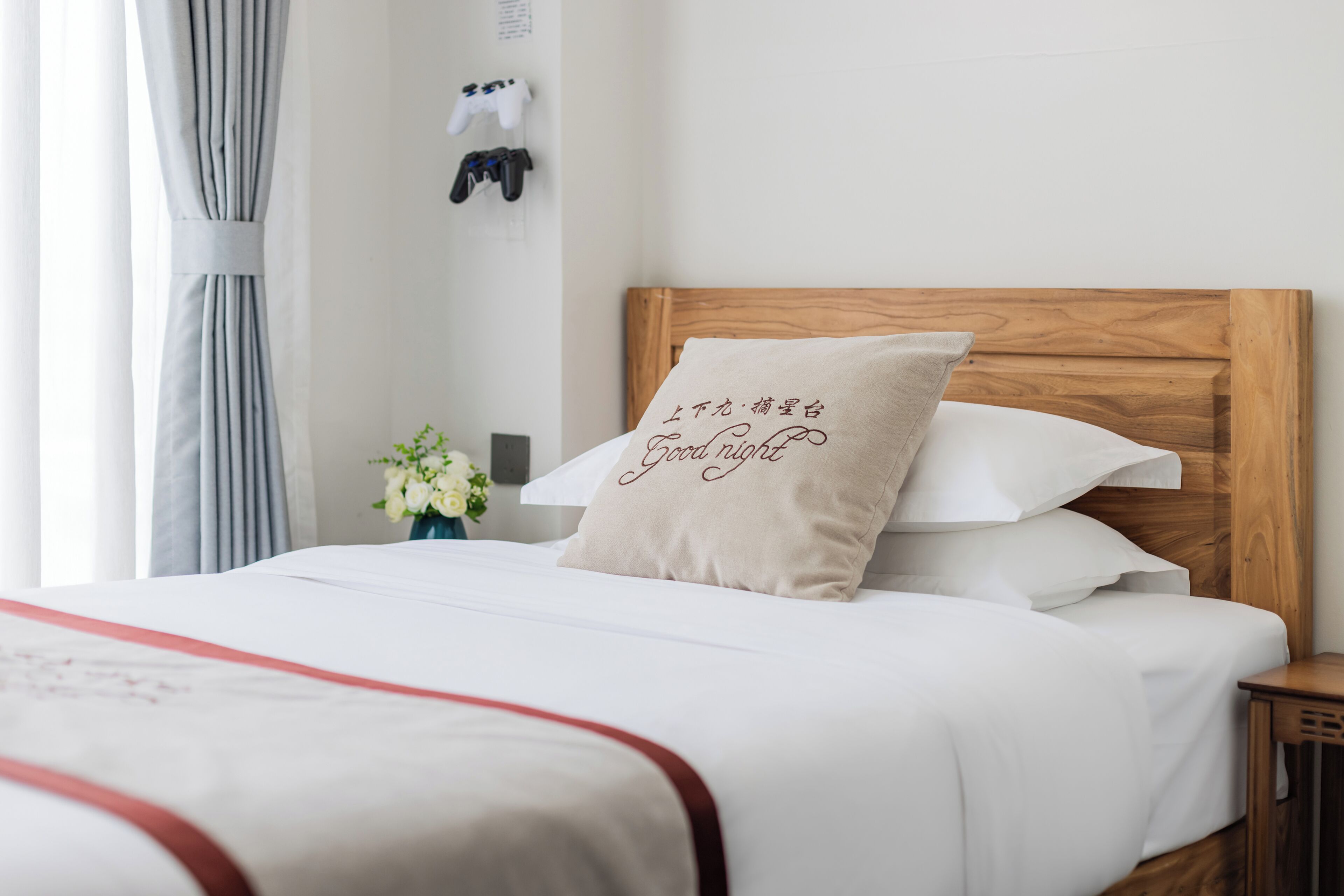 A neatly made bed with a wooden headboard, a pillow with text, and a small bedside table with a vase of flowers.