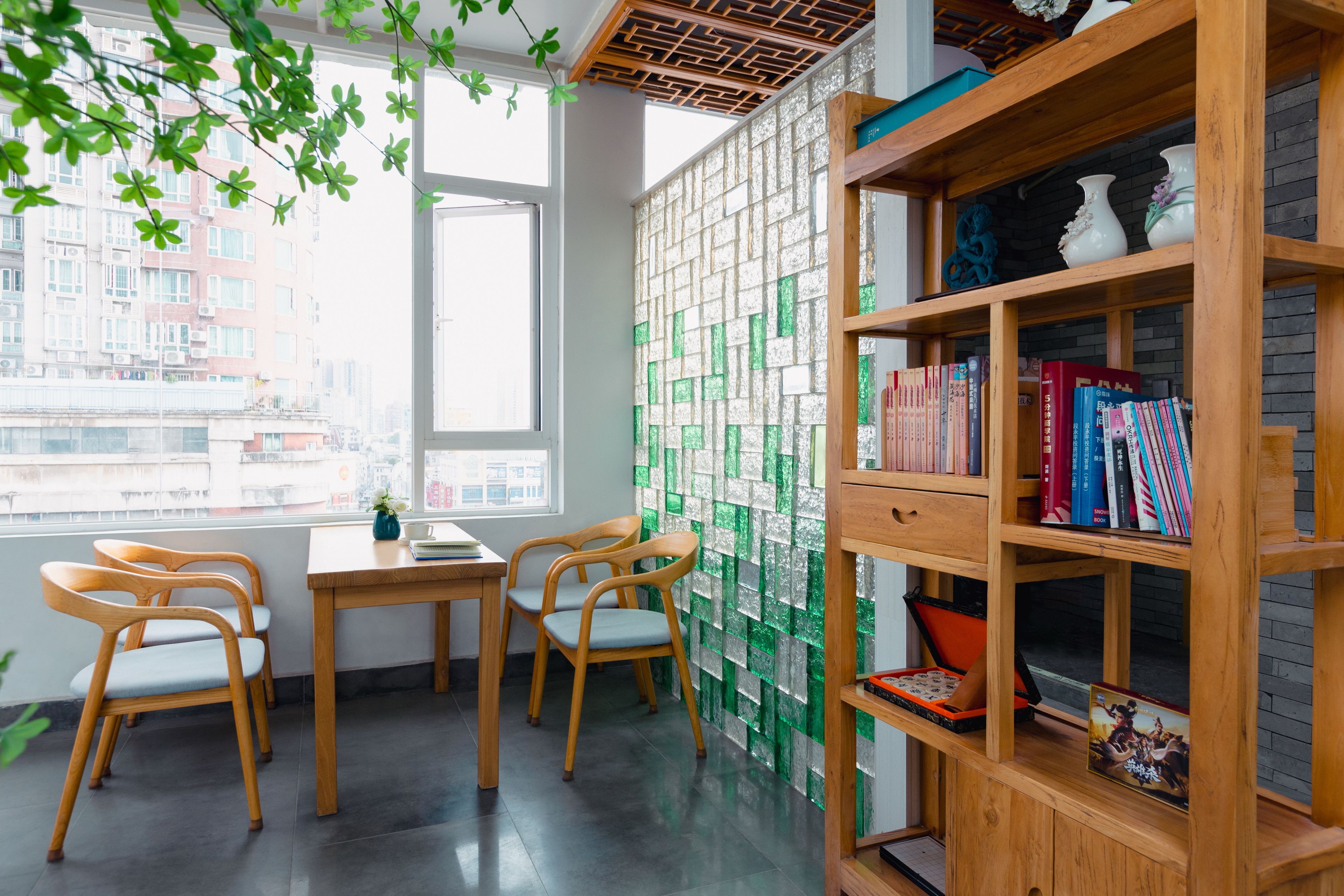 A room with a wooden desk, two chairs, a bookshelf, and a window with a view of buildings.