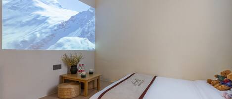 A bedroom with a bed, a bedside table with a vase and bottles, a projector screen displaying a snowy mountain scene, and a teddy bear on the bed.