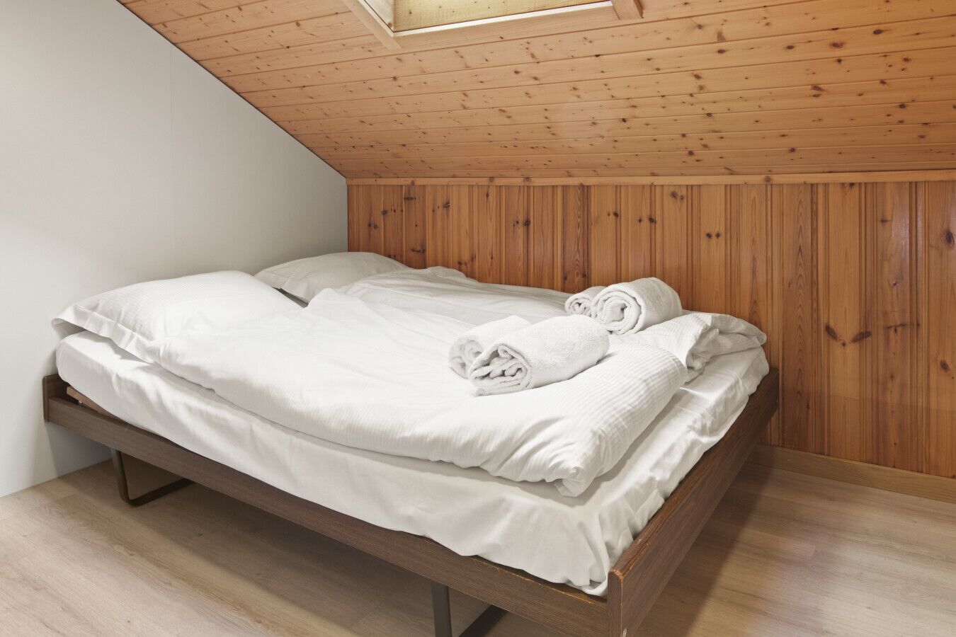 Een enkel bed met witte lakens en opgerolde handdoeken, een houten hoofdbord, en een schuine plafond met houten balken.