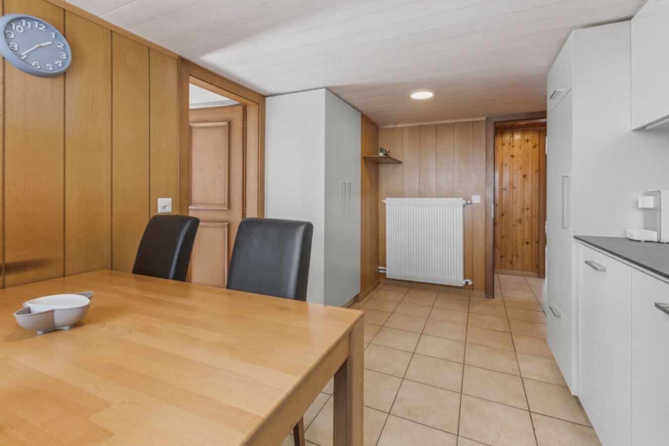 A compact kitchen with a wooden table, chairs, a clock, and white cabinets.