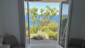 Un dormitorio con cama, mesita de noche, una silla y vista al océano a través de una puerta abierta.