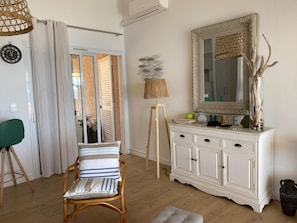 Una habitación con un armario blanco, un espejo, una silla de mimbre, una lámpara y vistas a través de una puerta.