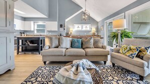 A spacious living area with a high ceiling, a kitchen in the background, and a large window providing natural light.
