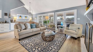 A spacious living room with a large sofa, a round coffee table, and a patterned rug. There is a dining area with a table and chairs, and a kitchen in the background.