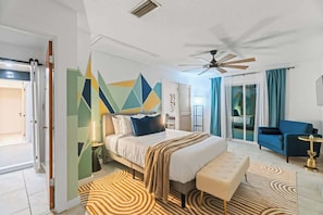 Une chambre d’hôtel moderne, équipée d’un lit, d’un ventilateur de plafond, d’un fauteuil bleu, d’une petite table ronde et offrant une vue sur l’extérieur.