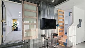 Een modern interieur met een aan de muur bevestigde televisie, twee barkrukken, een trap met houten treden en een metalen leuning.