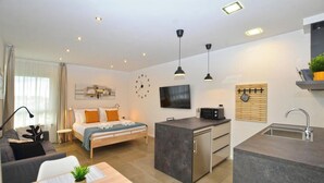 Un espace de vie moderne et compact comprenant un lit, un petit coin cuisine, une table à manger et un téléviseur fixé au mur.