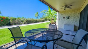A covered patio with a round table and chairs, a ceiling fan, and a view of a landscaped garden with palm trees and red flowers.