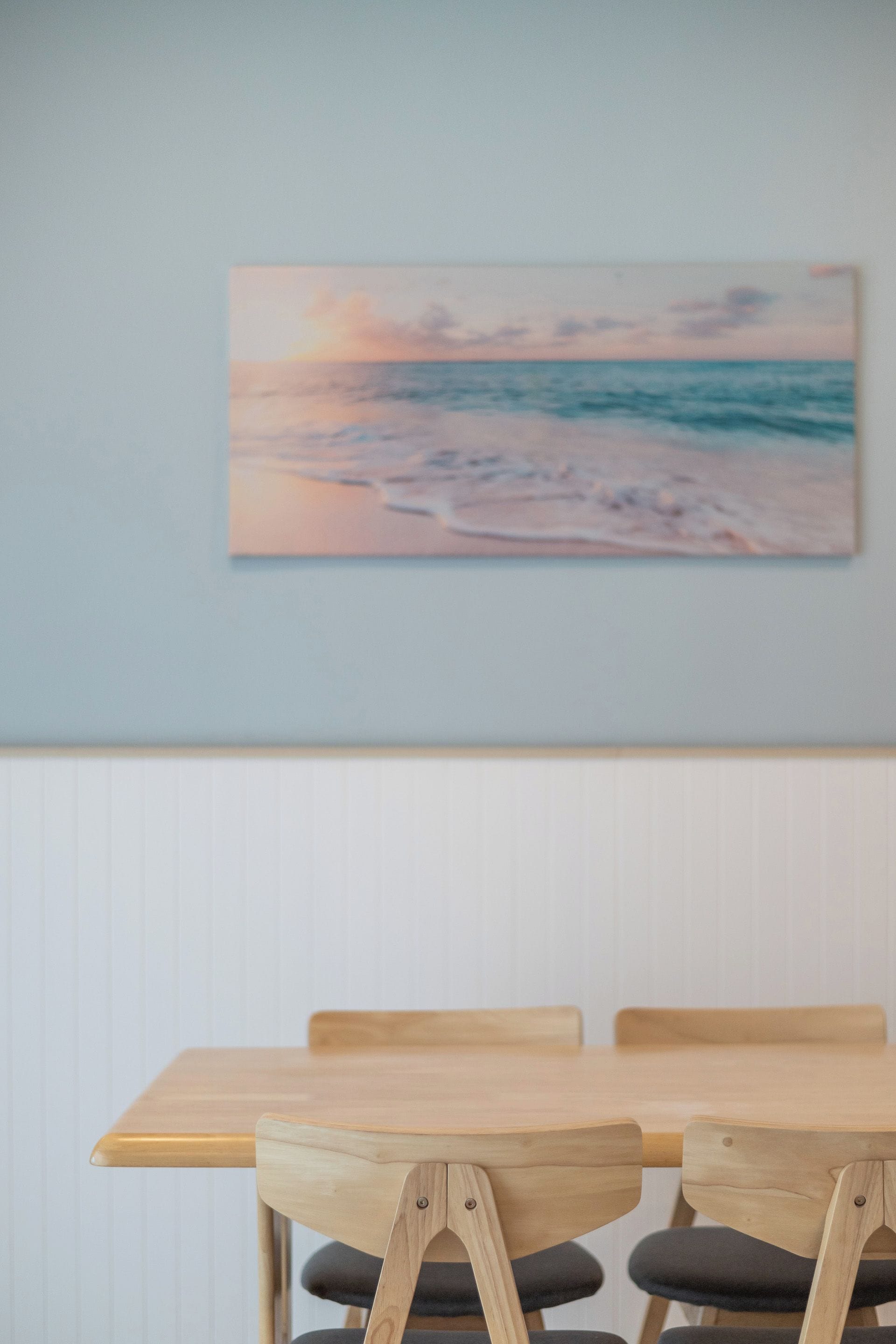 Un coin repas avec une table et des chaises en bois, un tableau représentant une scène de plage accroché au mur, et un mur blanc avec des panneaux décoratifs.